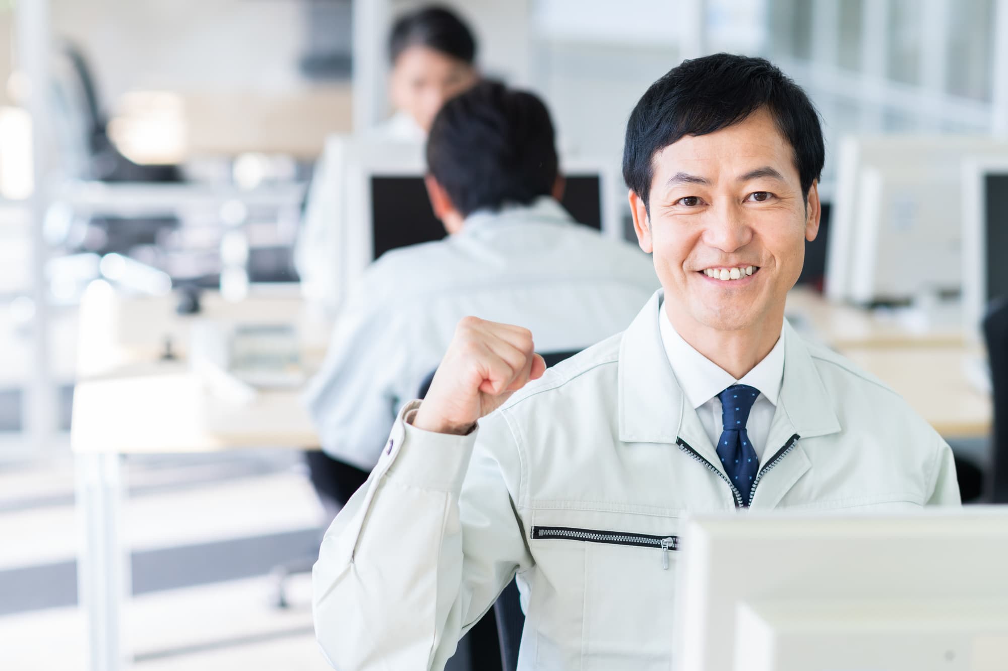 明るい事務所内にて品良くガッツポーズをする作業着の男性と、その背後で仕事に取り組む社員達